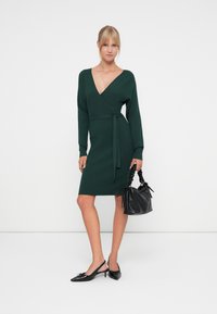 Robe tricotée verte à côtes avec un design enroulé, des manches longues et une ceinture à nouer. Portée avec des escarpins noirs à bride arrière et un sac à main noir texturé.