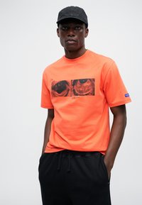 Tricou din bumbac portocaliu, cu un grafic rectangular ce are ilustrații detaliate ale ochilor. Capac negru purtat cu pantaloni negri cu șnur.