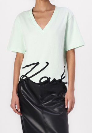 Camiseta de algodón verde menta con escote en V y mangas cortas, con un acento bordado en negro con la letra "K" en el dobladillo, combinada con una falda de cuero negra.