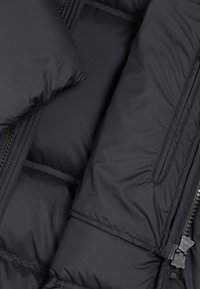 Chaqueta acolchada negra con cremallera visible y bolsillo cosido en tela lisa y aislante, en primer plano.