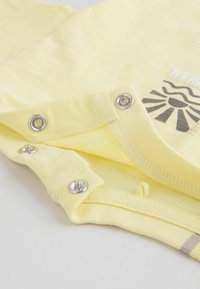 Vêtement pour bébé jaune avec des boutons-pression argentés le long de la couture et un graphisme partiel de soleil noir sur un tissu doux.