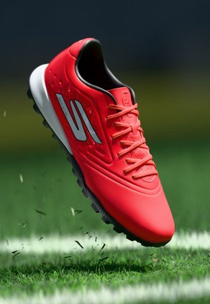 Lys rød fotballsko med svart såle, lisser bundet, svevende over en gressmatte med hvit grense og flygende gressstrå.