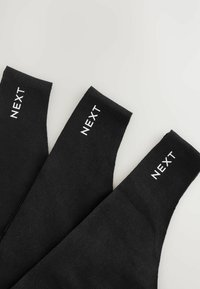 Tres calcetines negros apilados, cada uno exhibiendo el nombre de la marca "NEXT" en letras blancas en la banda superior. Fondo brillante.