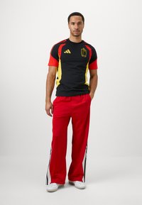 Sort og rød atletisk skjorte med gule detaljer, der har et logo. Parret med løstsiddende røde bukser, der fremviser hvide og sorte sidestriber.