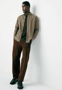 Man leaning against white wall wearing a brown plaid shirt over dark brown top, brown pants, and black shoes with hands in pockets.
