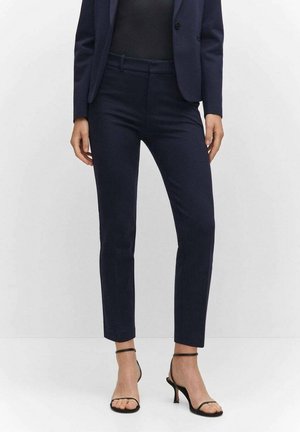 Vrouw die marineblauwe, nauwsluitende enkelbroeken draagt, bijpassend colbert, zwart topje en zwarte sandalen met hoge hak en bandjes, tegen een witte achtergrond.
