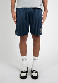 Homme portant un short de basket rayé bleu marine, des chaussettes blanches mi-mollet avec un texte, des mocassins noir et blanc, et un t-shirt gris clair.