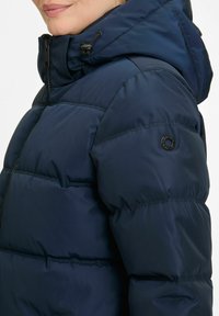 Vrouw in een marineblauwe gewatteerde jas met een hoge kraag en capuchon, dichtgeritst, met een close-up van de schouder en de bovenarm.
