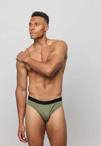 Grüne Baumwollunterhosen mit einem schwarzen Bündchen, die ein niedriges Design und eine glatte Textur aufweisen. Das Modell posiert mit einem Arm auf seiner Schulter.