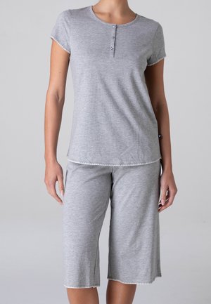 Donna che indossa un top a maniche corte grigio chiaro con colletto con bottoni e pantaloni larghi coordinati fino al ginocchio su sfondo semplice.