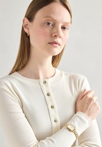 Cream ribbed long-sleeve top with five button placket. Model wears a gold watch with a square face and chain link band.