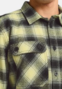 Chemise en flanelle à carreaux jaune et noir avec des boutons noirs proéminents, un col et une poche poitrine avec fermeture à bouton.