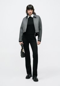 Sivá skrátená bunda so zipsom, čierny sveter a čierne nohavice. Modelka drží čiernu kabelku a nosí mohutné čierne topánky.