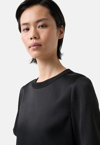 Černé tričko s krátkým rukávem, hladkým povrchem, kulatým výstřihem a uvolněným střihem. Tkanina se zdá být lehká a elegantní.