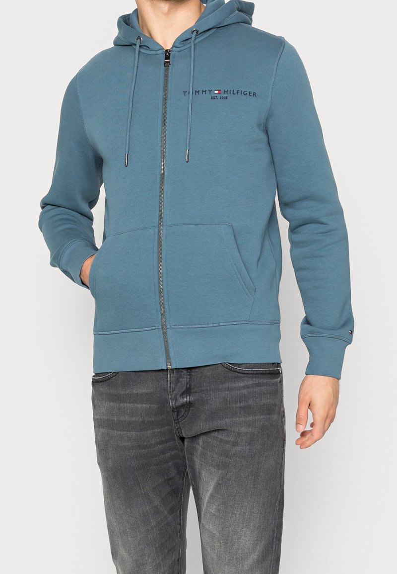 Sudadera azul con cremallera hecha de tela suave, con un logo en la parte frontal, dos bolsillos y capucha con cordón. Usada con jeans gris oscuro.
