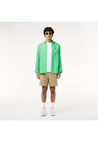 Camicia verde a bottoni, t-shirt bianca, pantaloni corti beige e sneakers bianche. Presenta una tasca sul petto e occhiali da sole tondini. Sfondo semplice.