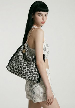 Jeune femme aux longs cheveux noirs portant un crop top fleuri et un short, portant un grand sac à épaule noir et blanc à motif.