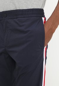 Navy blue athletic pants with an elastic waistband, featuring white and red side stripes and a side pocket. Textured, lightweight material.