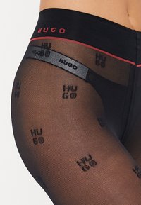 Juodos tinklinės pėdkelnės su raudonu HUGO logotipu ant juosmens ir pakartotiniu juodu "HUGO" prekės ženklu visame paviršiuje. Lygus paviršius su permatomu dizainu.