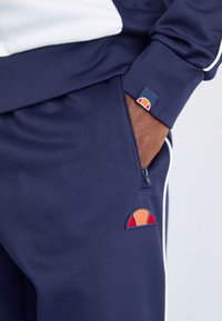 Marine trainingspak met witte accenten, featuring een ritszak. Opvallende geborduurde graphic in warme kleuren op de zak. Zachte textuur.