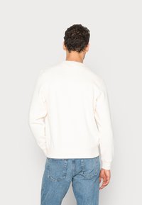 Sudadera de cuello redondo de color crema con mangas largas, puños y dobladillo de canalé, llevada con unos vaqueros de mezclilla de color azul claro. Textura de tela suave, diseño minimalista.