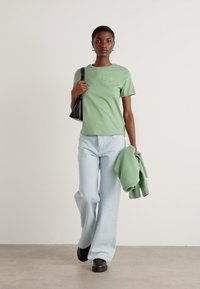 T-shirt verde menta, jeans svasati in denim chiaro, borsa nera, maglione verde chiaro e scarpe nere; tessuto morbido e vestibilità rilassata.
