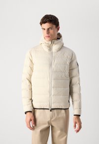 Jonge man in een lichtbeige gewatteerde jas met capuchon, staande met de handen ontspannen langs zijn zij, tegen een effen grijze achtergrond.