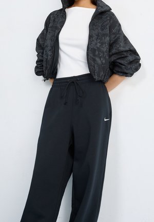 Femme portant un haut blanc ajusté, une veste courte noire à motif floral avec manches bouffantes, et un pantalon noir Nike à cordon de serrage.