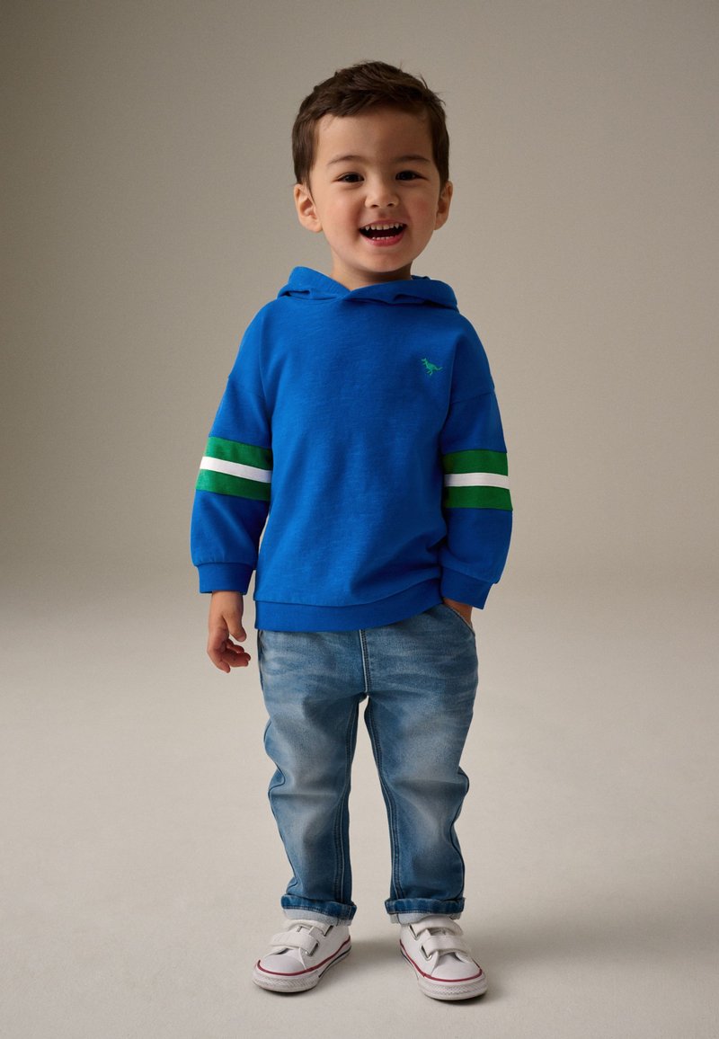 Ragazzo giovane in piedi che sorride, indossa una felpa con cappuccio blu con strisce verdi e bianche, jeans azzurri chiari e scarpe da ginnastica bianche.