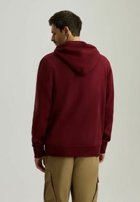 Bordowy hoodie z kapturem na sznurki, ribbowymi mankietami i dołem. Tkanina wygląda na miękką, o prostym designie, noszona na khaki spodniach.