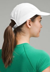 Witte pet met een gebogen klep, gaasstructuur en verstelbare achterkant. Het onderwerp draagt een groene sportshirt en heeft het haar in een paardenstaart.