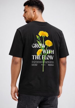 Man wearing black t-shirt with yellow dandelion graphic and white text "Grow with the flow" on the back against a plain background.