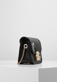 Černá kožená kabelka se zlatým řetízkem, zlatým uzávěrem a bílou květinovou výšivkou na předním klopě ležící na bílé ploše.