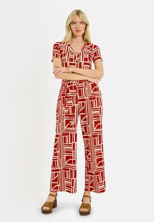 Femme portant une combinaison courte à manches courtes avec un motif géométrique rouge et blanc, avec des boutons, une ceinture, des sandales à plateforme beiges, des lunettes et un bracelet, debout.