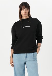 Zwarte sweatshirt met een ronde hals en lange mouwen, voorzien van het witte "Calvin Klein" logo aan de voorkant. Draagt met lichtblauwe jeans.