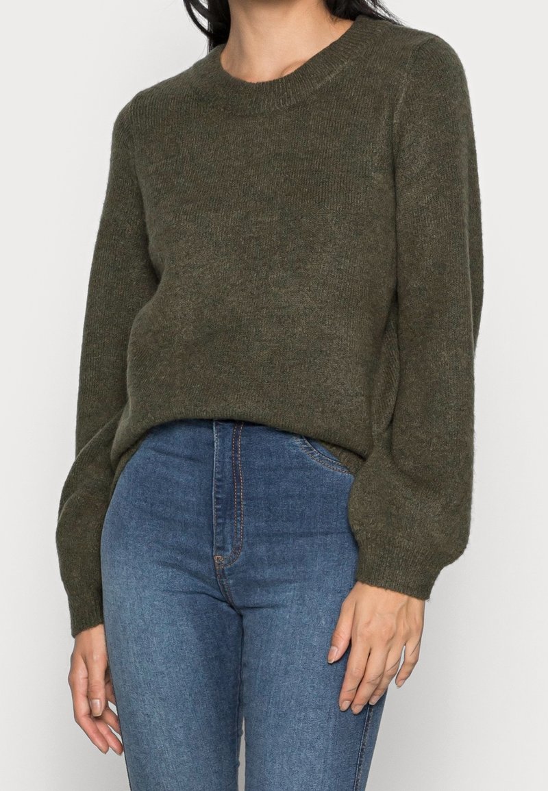 Femme portant un pull en maille vert olive à manches longues avec des poignets côtelés, associé à un jean bleu taille haute, vue de la taille vers le haut.