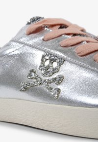 Zapatilla metálica plateada que presenta un diseño brillante de calavera y huesos cruzados. Los cordones en un suave tono rosa y la suela blanca texturizada completan el look.