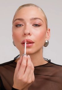 Frau mit nach hinten gegelten Haaren, die rosa Lipgloss aufträgt, trägt silberne Ohrringe und ein schwarzes durchsichtiges Oberteil vor hellem Hintergrund.