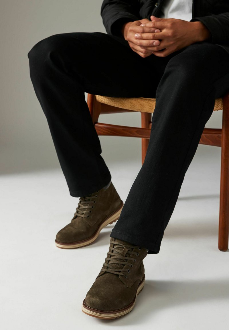 Botas de tobillo de ante marrón con diseño de cordones, suela de goma y detalles en color beige. Llevadas con pantalones negros mientras se está sentado en una silla de madera.