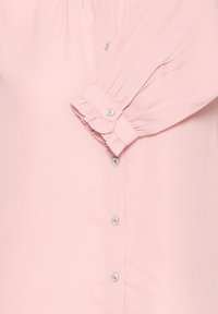 Lichtroze blouse mouw met gerimpelde manchet en parelachtige knoop, met een deel van de geknoopte voorkant zichtbaar.
