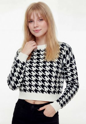 Noor naine sirgete blondide juustega, kes kannab mustvalget koerahammasmustriga kampsunit ja musti p ükse, poseerides ühe käega kaela lähedal.