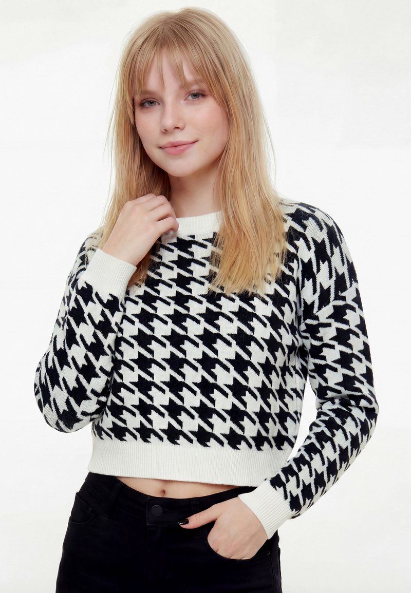 Mladá žena s rovnými blond vlasmi, oblečená v čierno-bielom svetri so vzorom psích zubov a čiernych nohaviciach, pózujúca s jednou rukou pri krku.