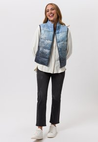 Blauwe gewatteerde bodywarmer met een verloopmotief, rits aan de voorkant en hoge kraag. Gecombineerd met een wit overhemd, zwarte spijkerbroek en witte sneakers.