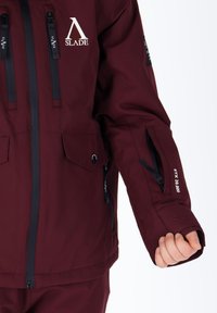 Giacca impermeabile color marrone bordeaux con logo, tasche con zip, polsini regolabili e finitura in tessuto con un aspetto liscio.