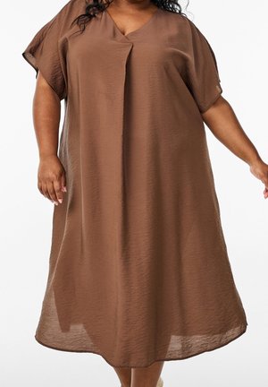 Femme portant une robe marron ample, longueur genou, avec des manches courtes et un col en V, debout les bras légèrement étendus.