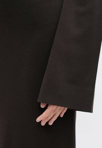 Długi rękaw w ciemno brązowej teksturowanej tkaninie, z szerokim, prostym rękawem i dłonią opartą na tkaninie.