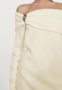 Pull léger crème à épaules dénudées avec une texture douce, comportant un bijou décoratif en argent à l'épaule. Tissu lisse et drapé.