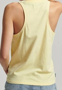 Top de algodón amarillo sin mangas con cuello redondo y diseño de espalda abierta, que presenta un dobladillo cosido y una marca mínima en la parte inferior derecha.
