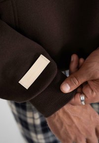 Primer plano de las manos de una persona llevando un anillo de plata, ajustando el puño de una sudadera marrón oscuro con una etiqueta beige que dice "PROSHIFTED".