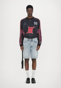 Schwarzes Langarmtrikot mit lila Akzenten und Grafiken, kombiniert mit hellblauen Denim-Shorts, weißen Socken und schwarzen Slippern.